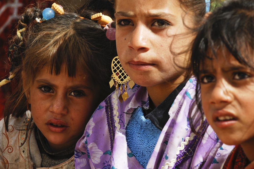 A close-up photograph of three young girls with serious or concerned expressions on their faces. The central figure wears decorative gold earrings and a purple floral garment, while the girl on the left has colorful hair accessories. The image has a documentary or photojournalistic quality.