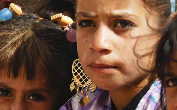 A close-up photograph of three young girls with serious or concerned expressions on their faces. The central figure wears decorative gold earrings and a purple floral garment, while the girl on the left has colorful hair accessories. The image has a documentary or photojournalistic quality.
