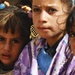 A close-up photograph of three young girls with serious or concerned expressions on their faces. The central figure wears decorative gold earrings and a purple floral garment, while the girl on the left has colorful hair accessories. The image has a documentary or photojournalistic quality.