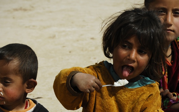Three young children are seated on sandy ground eating from metal bowls and plates. The central child, wearing an orange/mustard garment, is holding a spoon with white food (likely rice) toward their mouth with tongue extended. The children appear to be in an outdoor, arid environment with minimal resources visible.