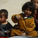 Three young children are seated on sandy ground eating from metal bowls and plates. The central child, wearing an orange/mustard garment, is holding a spoon with white food (likely rice) toward their mouth with tongue extended. The children appear to be in an outdoor, arid environment with minimal resources visible.