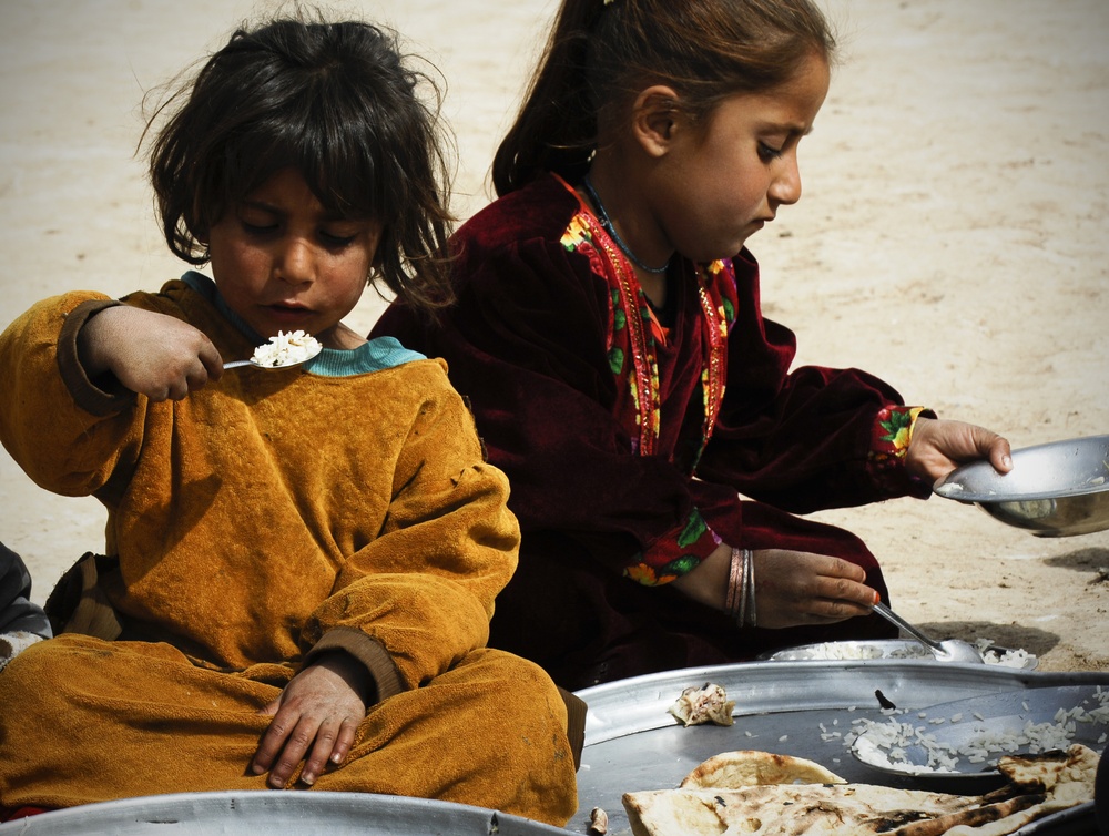 Two young children are seated on sandy ground eating a meal from large metal platters. The younger child on the left wears an orange outfit and holds a spoon with rice, while the older child on the right wears a dark red embroidered garment. The scene suggests a modest outdoor meal setting, likely in a Central or South Asian context based on the clothing styles.