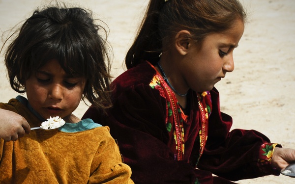 Two young children are seated on sandy ground eating a meal from large metal platters. The younger child on the left wears an orange outfit and holds a spoon with rice, while the older child on the right wears a dark red embroidered garment. The scene suggests a modest outdoor meal setting, likely in a Central or South Asian context based on the clothing styles.