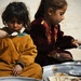 Two young children are seated on sandy ground eating a meal from large metal platters. The younger child on the left wears an orange outfit and holds a spoon with rice, while the older child on the right wears a dark red embroidered garment. The scene suggests a modest outdoor meal setting, likely in a Central or South Asian context based on the clothing styles.