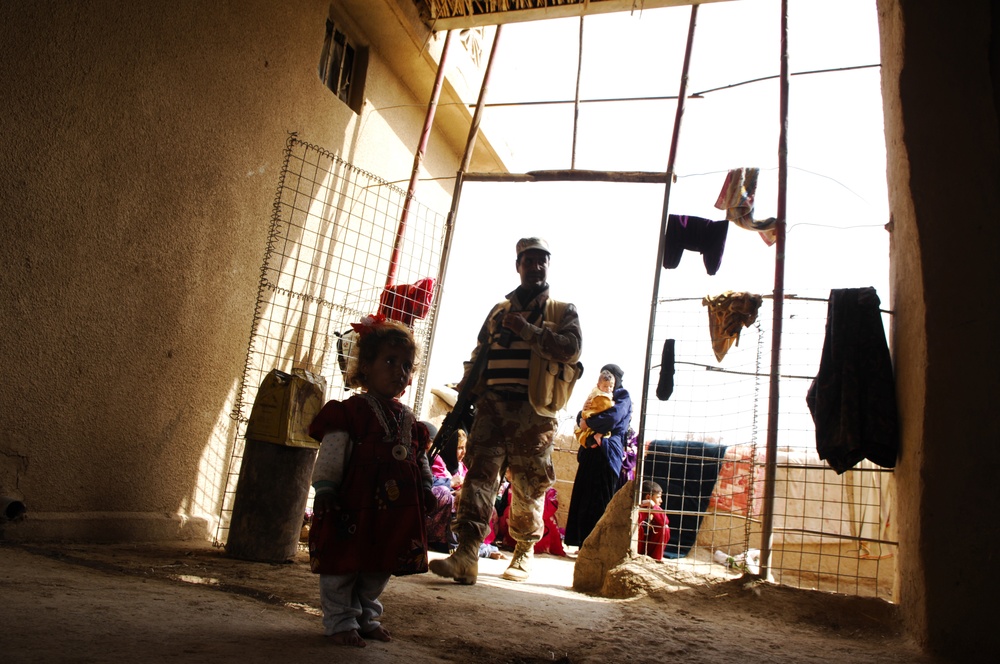 The image shows a dimly lit interior space, likely a courtyard or transitional area of a modest dwelling, with a uniformed soldier standing in a bright doorway. Several children and at least one adult woman are visible in the background. Clothing items are hanging on lines strung across the entrance frame.