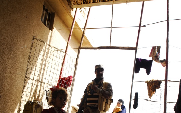 The image shows a dimly lit interior space, likely a courtyard or transitional area of a modest dwelling, with a uniformed soldier standing in a bright doorway. Several children and at least one adult woman are visible in the background. Clothing items are hanging on lines strung across the entrance frame.