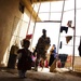 The image shows a dimly lit interior space, likely a courtyard or transitional area of a modest dwelling, with a uniformed soldier standing in a bright doorway. Several children and at least one adult woman are visible in the background. Clothing items are hanging on lines strung across the entrance frame.
