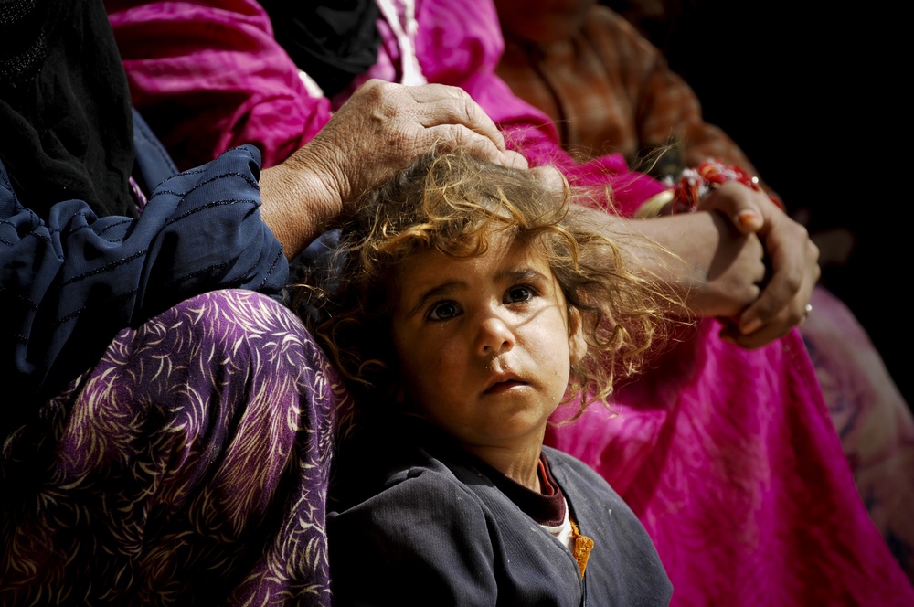 A young child with curly, disheveled hair looks upward with a solemn, wide-eyed expression. An adult hand rests on the child's head, suggesting a protective or comforting gesture. The child is surrounded by adults wearing colorful traditional garments in magenta, purple, and dark blue.