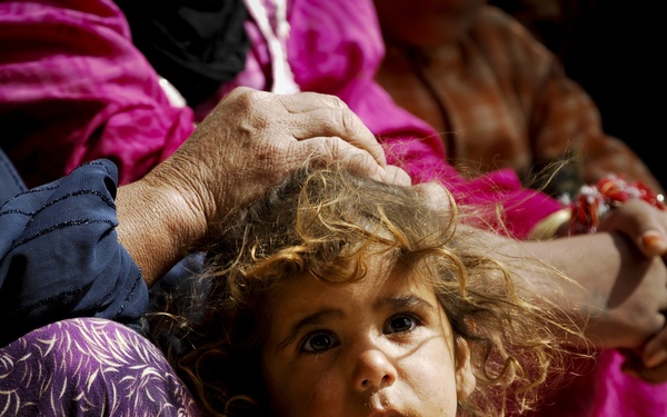 A young child with curly, disheveled hair looks upward with a solemn, wide-eyed expression. An adult hand rests on the child's head, suggesting a protective or comforting gesture. The child is surrounded by adults wearing colorful traditional garments in magenta, purple, and dark blue.