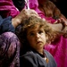 A young child with curly, disheveled hair looks upward with a solemn, wide-eyed expression. An adult hand rests on the child's head, suggesting a protective or comforting gesture. The child is surrounded by adults wearing colorful traditional garments in magenta, purple, and dark blue.