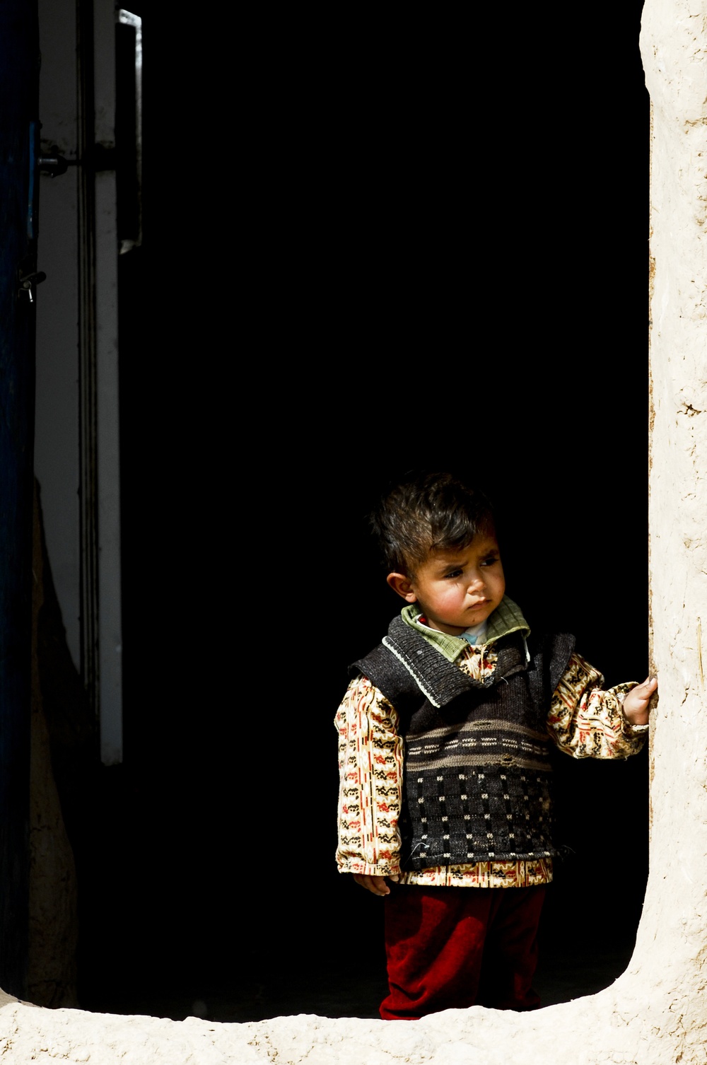 A young toddler stands in a doorway or threshold of a building with whitewashed walls, leaning slightly against the door frame. The child is dressed in multiple layered garments including a patterned long-sleeve shirt, a dark knitted vest, and red trousers. The interior behind the child is very dark, creating a strong contrast with the sunlit exterior wall.