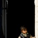 A young toddler stands in a doorway or threshold of a building with whitewashed walls, leaning slightly against the door frame. The child is dressed in multiple layered garments including a patterned long-sleeve shirt, a dark knitted vest, and red trousers. The interior behind the child is very dark, creating a strong contrast with the sunlit exterior wall.