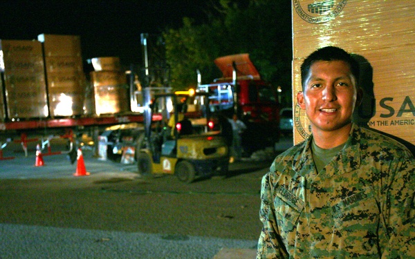 A U.S. Marine in digital camouflage uniform stands beside stacked USAID boxes during a nighttime humanitarian aid distribution operation. Forklifts and flatbed trucks loaded with USAID-branded cargo are visible in the background, indicating an active logistics operation. The scene depicts a military-supported USAID relief effort, likely in response to a disaster or humanitarian crisis.