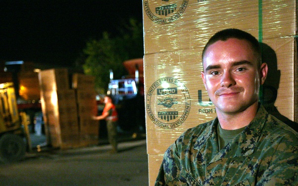 A U.S. Marine in digital camouflage (MARPAT) uniform stands with arms crossed in front of stacked USAID (United States Agency for International Development) boxes at night. The setting appears to be a humanitarian aid distribution or logistics operation, with forklifts and workers visible in the background. The boxes are marked with USAID branding, indicating international development or disaster relief supplies.