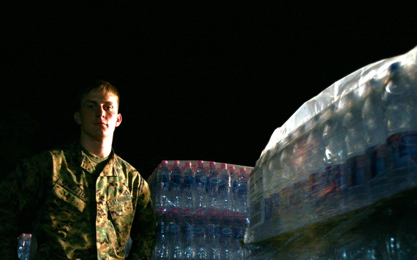 A military service member in camouflage uniform stands among large pallets of bottled water wrapped in plastic shrink wrap, photographed at night. The scene appears to be a humanitarian aid or logistics operation involving water distribution. The soldier holds a single water bottle and is illuminated against a dark background.