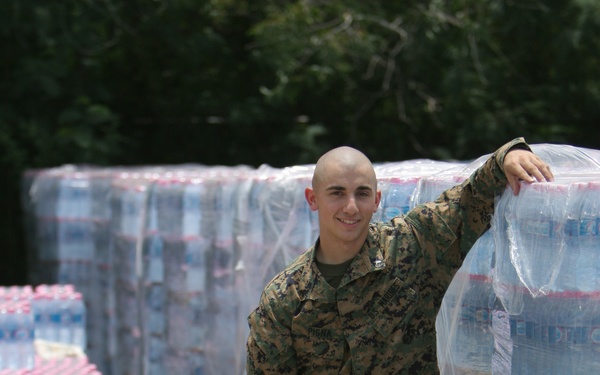 A young male service member in U.S. Marine Corps MARPAT digital camouflage uniform is posing beside pallets of shrink-wrapped bottled water. He is smiling and leaning casually against one of the pallets, suggesting a humanitarian aid or disaster relief supply operation. Multiple pallets stacked with water bottles are visible in the background.