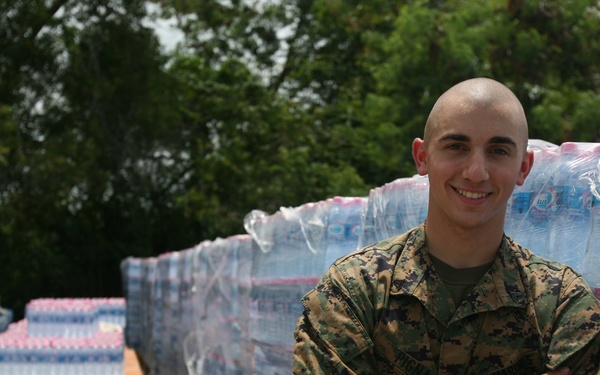 A young male service member in U.S. Marine Corps MARPAT camouflage uniform stands with arms crossed and smiling in front of large pallets of bottled water wrapped in plastic. The setting appears to be an outdoor staging or distribution area, likely associated with disaster relief or humanitarian aid operations. Multiple pallets of water bottles extend into the background alongside additional stacked cases.