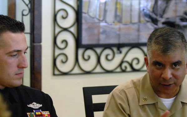 Two U.S. Navy personnel are seated at a dining table during what appears to be a formal or semi-formal military luncheon or dinner event. The senior officer on the right, whose name tag reads 'JOE CAMP[...], is speaking and gesturing, while the junior enlisted sailor on the left listens attentively. The setting includes a decorated table with beverages, food, and a carved watermelon centerpiece.