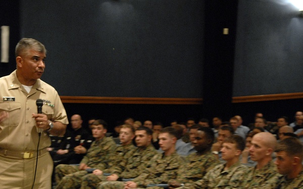 A senior military officer in tan/khaki Navy service uniform is addressing a large group of seated military personnel, predominantly in Marine Corps MARPAT camouflage uniforms. The speaker is holding a microphone and gesturing with his free hand, suggesting an all-hands briefing or command presentation. The event appears to take place in an auditorium or theater-style venue.