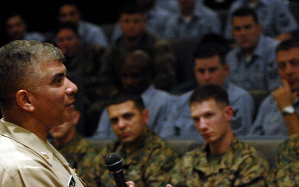A senior military officer in a tan/khaki dress uniform is addressing a large audience of uniformed service members in what appears to be an auditorium or theater setting. The speaker is holding a microphone and gesturing with both hands, suggesting an active briefing or all-hands meeting. The audience, wearing Marine Corps MARPAT camouflage uniforms, is attentive and seated in rows.
