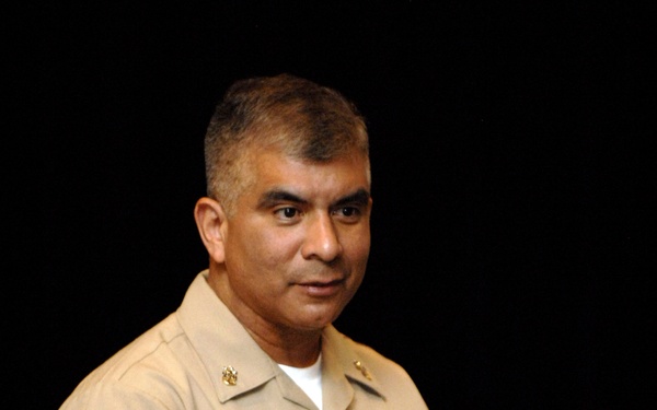 A U.S. Navy enlisted sailor named Joe Campa is photographed in khaki service uniform while speaking or presenting, gesturing with both hands. He is wearing a name tag reading 'JOE CAMPA,' service ribbons, a warfare device badge, and a watch and ring. The background is dark with a hint of wooden paneling, suggesting an auditorium or formal meeting setting.