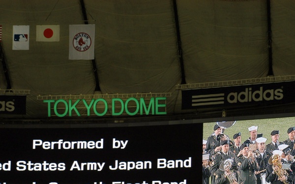 The image shows the scoreboard at Tokyo Dome during a baseball event, displaying a performance announcement for the United States Army Japan Band and U.S. Navy's Seventh Fleet Band. The venue is hosting what appears to be a Major League Baseball international game, evidenced by the Oakland Athletics and Boston Red Sox pennants alongside the American and Japanese flags. A video screen shows uniformed military band members performing.