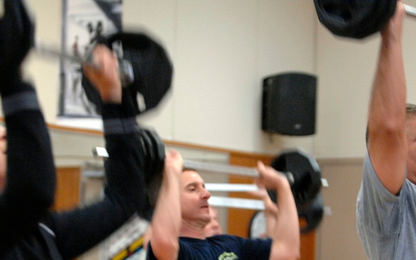 Three individuals are performing overhead barbell lifts in a gym setting. The group includes a woman on the left in black athletic wear and two men, all pressing barbells loaded with weight plates above their heads. The setting appears to be a fitness facility with equipment and motivational posters visible in the background.