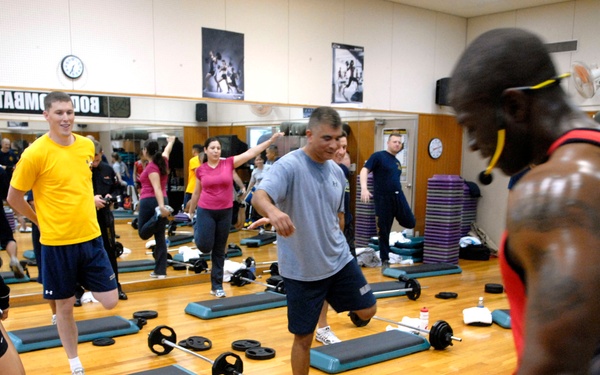 A group fitness class is taking place in a gymnasium or fitness center, with participants performing step aerobics or a combat fitness routine using step platforms and barbells with weight plates. The class appears to be a structured group exercise session, possibly a BodyCombat or similar program as indicated by signage on the wall. Multiple participants of varying ages and fitness levels are engaged in lunging or stepping movements around the equipment.