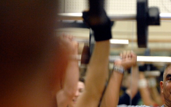 A group of people are exercising in a gym, performing overhead barbell or weight lifts. The most prominent figure in focus is a middle-aged man wearing a gray Under Armour shirt, lifting a barbell overhead. Several other participants, including a woman in a pink top, are visible in the background performing similar exercises.