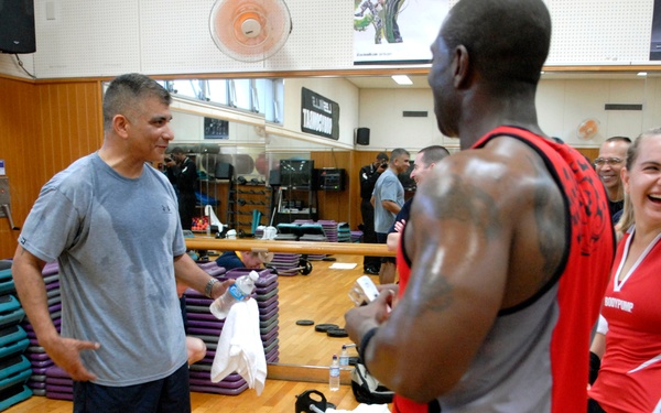 A group of people are gathered in a gymnasium or fitness facility, engaged in a social interaction after or during a workout session. A muscular man in a red tank top is conversing with a man in a gray t-shirt, while others nearby laugh and smile. Weight plates, barbells, and exercise mats are visible on the floor, indicating a group fitness class setting.