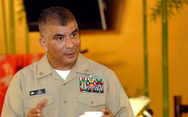 Two U.S. Navy personnel in khaki service uniforms are engaged in conversation at what appears to be a formal dining or luncheon event. The standing officer, identified by his name tag as 'Joe Campa,' holds a white coffee mug and gestures while speaking to a seated colleague. Both individuals display extensive ribbon/medal arrays and military insignia on their uniforms.