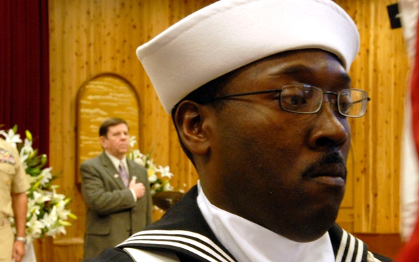A formal military ceremony is taking place in what appears to be a chapel or ceremonial hall. A U.S. Navy enlisted sailor in dress blues uniform with a white Dixie cup hat stands in the foreground holding a flag, while a naval officer in khaki uniform salutes in the background and a civilian in a suit stands with hand over heart. The setting suggests a patriotic ceremony such as a change of command, retirement, or memorial service.