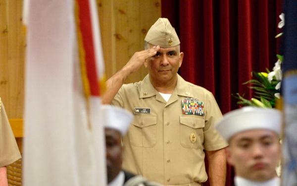 A formal U.S. Navy military ceremony is taking place indoors, likely a change of command or retirement ceremony. A senior Navy officer in the center is rendering a salute while a color guard of sailors in dress uniforms carries flags in the foreground. Additional uniformed personnel and a civilian in a suit are visible in the background.