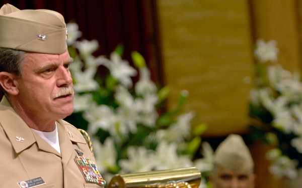 A senior military officer in tan/khaki service uniform stands at a wooden podium with a brass lamp, appearing to deliver a speech or address. A second uniformed military personnel is seated in the background. The setting appears to be a formal ceremony or chapel, decorated with white lily flower arrangements.