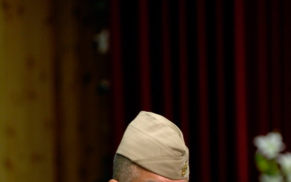 A U.S. Navy enlisted sailor identified by name tag as 'Joe Campa' stands at a wooden podium, appearing to address an audience at a formal military ceremony. He is wearing the tan/khaki Service Uniform (Service Khaki) with a garrison cover, multiple rows of service ribbons, a warfare device, and senior enlisted insignia. The setting appears to be an auditorium or ceremonial hall with red curtains and floral arrangements in the background.