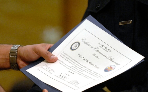 Two individuals are shaking hands during what appears to be a formal military or official ceremony. A certificate titled 'Certificate of Educational Achievement' from NAF ATSUGI, Japan is being presented, made out to 'YNC(AW/SW) Richard Harvey'. The certificate is held in a dark blue folder/portfolio during the handshake presentation.
