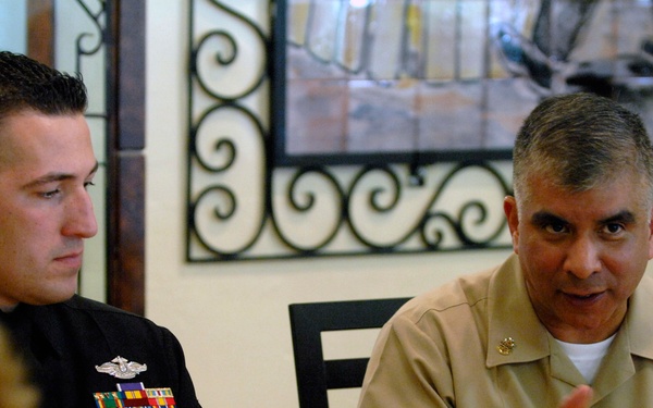 Two U.S. Navy personnel are seated at a dining table during what appears to be a formal or semi-formal military luncheon or dinner. The senior officer on the right, whose name tag reads 'JOE CAMP[...], is speaking and gesturing, while the junior enlisted sailor on the left listens attentively. The setting includes drinks, food, and decorative elements consistent with a restaurant or official dining facility.