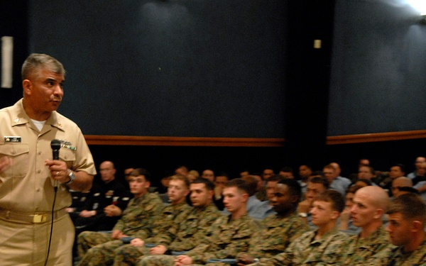A senior military officer in tan/khaki Navy service uniform is addressing a large group of seated military personnel, predominantly in Marine Corps MARPAT camouflage uniforms. The speaker is holding a microphone and gesturing with one hand, suggesting an all-hands briefing or command presentation. The event is taking place in what appears to be a military auditorium or theater.