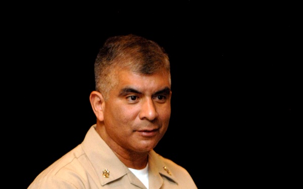 A U.S. Navy enlisted sailor named Joe Campa is photographed in khaki service uniform while speaking or presenting, gesturing with both hands. He is wearing a name tag reading 'JOE CAMPA,' service ribbons, a warfare device badge, and a watch and ring. The background is dark with a hint of wooden paneling, suggesting an auditorium or formal meeting setting.