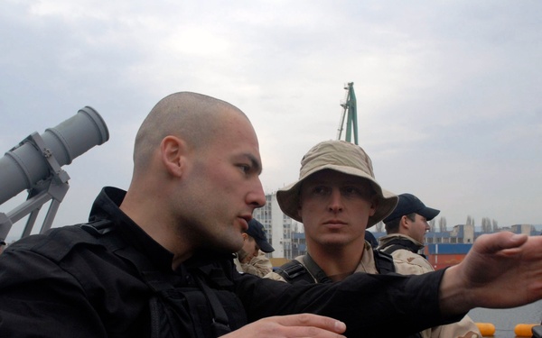 The image shows a group of military or security personnel on what appears to be the deck of a naval vessel in a harbor. A man in black tactical gear is gesturing with his hands, apparently briefing or instructing others wearing desert camouflage uniforms. Industrial port cranes and a cargo ship are visible in the background.