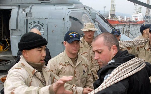 Military personnel in desert camouflage uniforms are gathered on the flight deck of a naval vessel, with a helicopter visible in the background. One service member in a black jacket is handling a thick rope while another in camouflage appears to be providing instruction or guidance. The scene appears to take place in a port environment, with cranes and a red ship visible in the background.