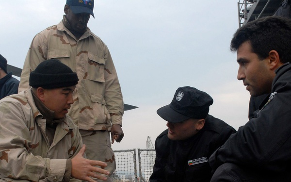 Military personnel in desert camouflage uniforms appear to be conducting a training or demonstration session on what appears to be a ship's deck. A crouching service member is showing or explaining something involving rope/cordage to two individuals dressed in black uniforms. The setting appears to be a naval vessel, likely during a joint training exercise.