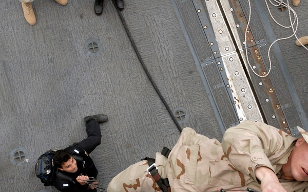 The image shows military personnel conducting a ship boarding or fast-rope/rappelling exercise on the exterior hull of a naval vessel. At least three individuals are visible at different stages of the descent or boarding operation. The personnel are wearing desert camouflage uniforms, suggesting a training or operational maritime security scenario.