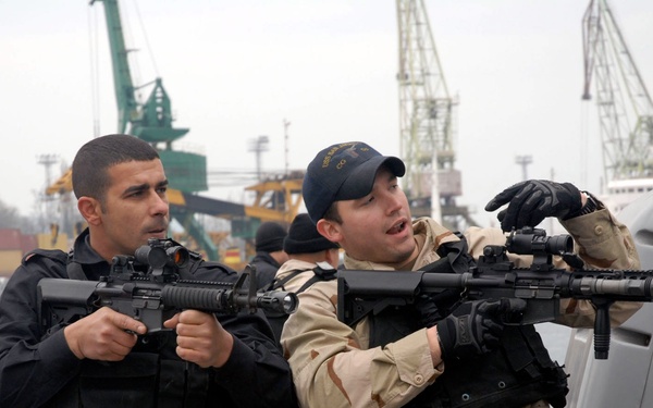 Two armed military or law enforcement personnel are positioned on what appears to be a naval vessel deck, holding assault rifles in a tactical stance. One individual wears all-black tactical gear while the other wears desert camouflage (DCU pattern) with a navy-style ball cap. The setting appears to be a port or harbor environment, suggesting a training exercise or security operation.