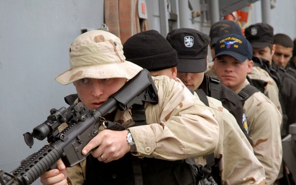The image shows military or security personnel aboard what appears to be a naval vessel, with the lead figure in desert camouflage uniform and boonie hat aiming an assault rifle. Multiple personnel are lined up behind the lead figure, wearing a mix of desert camouflage and dark tactical gear. A directional sign marked 'D' is visible on the gray bulkhead wall.