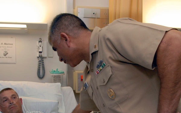 A military officer in tan/khaki uniform is leaning over and speaking with a patient lying in a hospital bed. The patient is wearing a hospital gown and has an IV line visible. A U.S. Marines sign is posted on the wall behind the bed, suggesting this is a military medical facility.