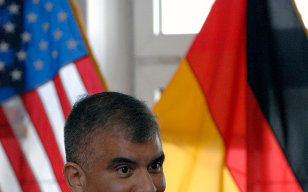 A U.S. Navy officer named Joe Campa is photographed speaking or briefing, with both hands raised in a gesturing position. He is wearing the tan/khaki Navy working uniform with a name tag, ribbons, and a warfare device badge. The background displays both a U.S. flag and a German flag, suggesting a bilateral or NATO-related military setting.
