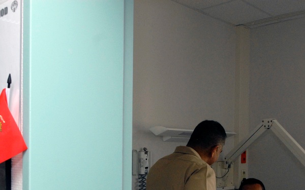 A hospital room scene showing a patient in bed using a laptop computer while a visitor or medical staff member stands beside the bed. The room is identified as Ward 10D, Bed #214, with the patient's name partially visible as 'L. H.' on a sign outside the door. Two small flags — an American flag and what appears to be a United States Marine Corps flag — are displayed on the door frame.
