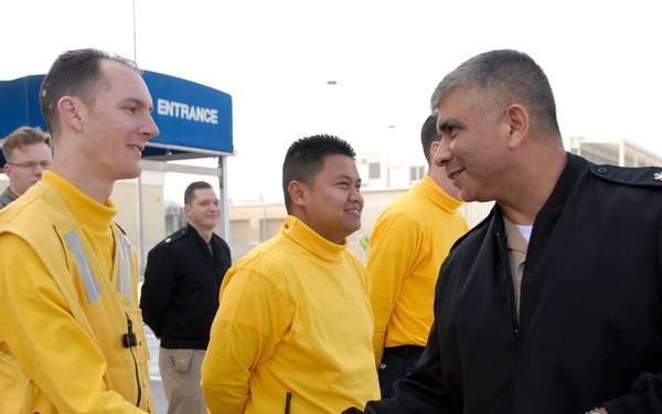 A senior military officer in a dark navy jacket and khaki trousers is shaking hands with a service member wearing a yellow flight deck jersey and navy trousers. Several other personnel in yellow jerseys are standing nearby, suggesting an official greeting or inspection visit. The setting appears to be a military installation entrance or flight line area.
