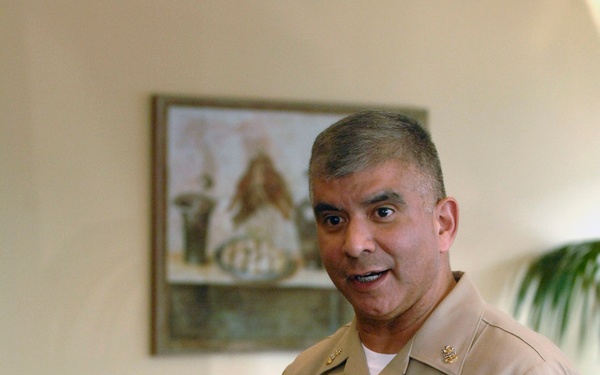 A U.S. Navy officer in khaki service uniform is speaking to a group, holding a white coffee mug and gesturing with his free hand. His name tag reads 'JOE CAMPA' and he wears several ribbons, a warfare qualification badge, and a large command/master chief insignia. The setting appears to be an informal meeting or gathering in a dining or conference room.
